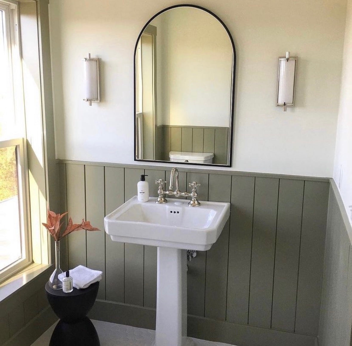 A white pedestal sink under an arched mirror in a sage-green bathroom